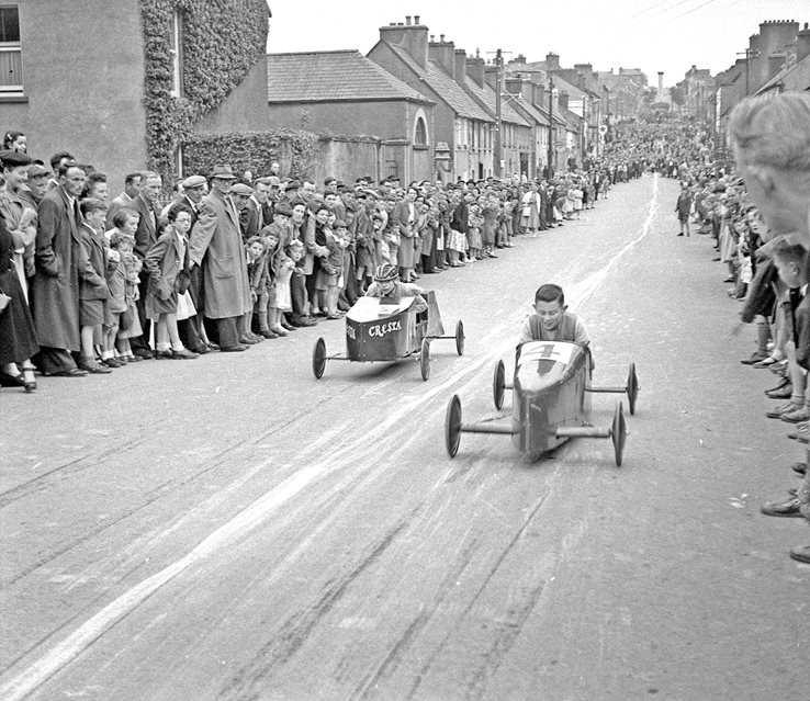 black and white photograph showing two soap box cars racing down a street with crowds watching from the sides.